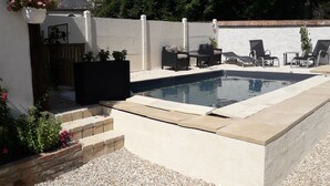 A rectangular swimming pool with a white border, surrounded by a white wall and a gravel area. There are several lounge chairs and a table near the pool.