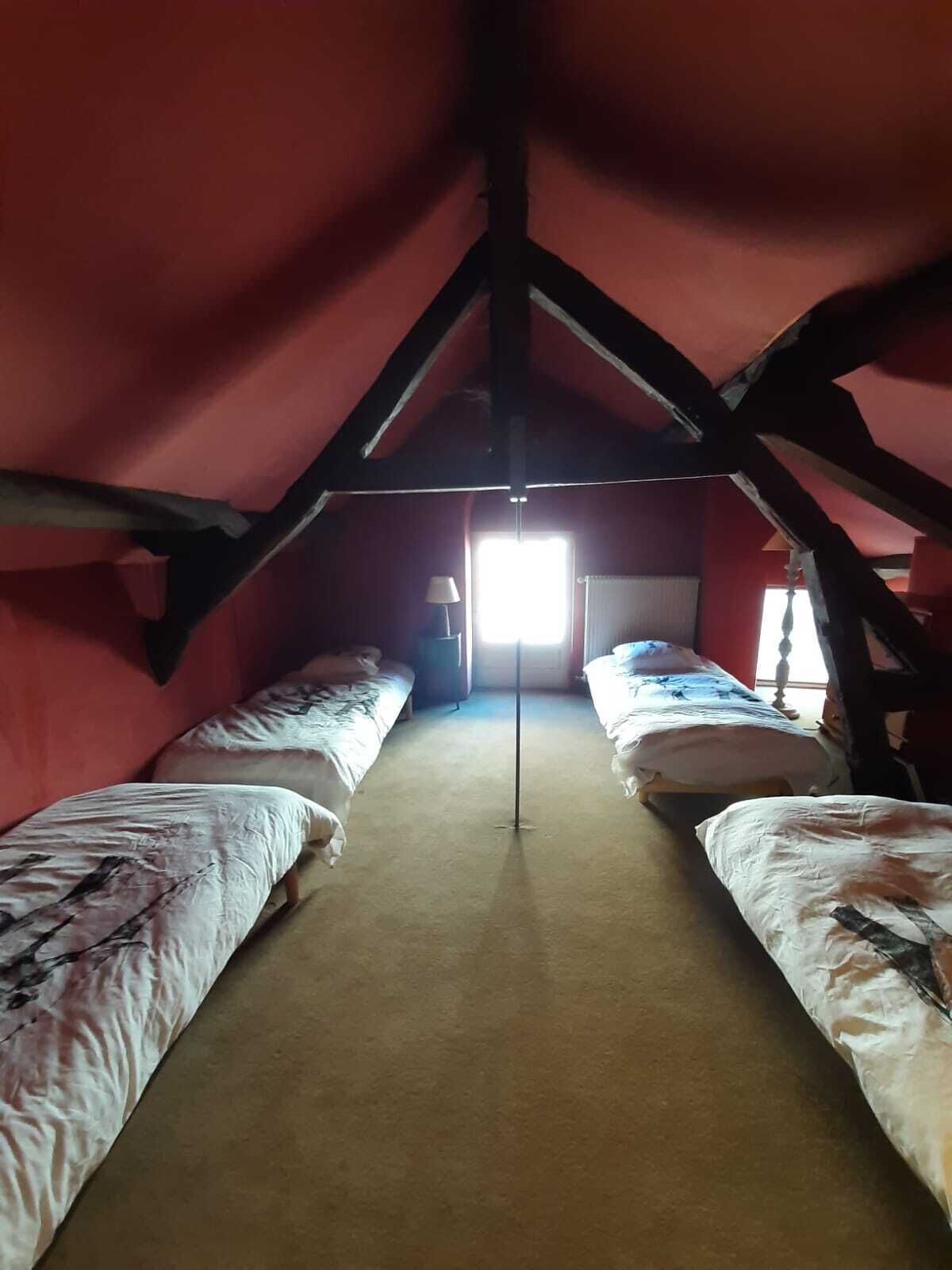 Habitación con dos camas dispuestas a lo largo de un eje central, una ventana que proporciona luz natural y una pared roja con techo inclinado.