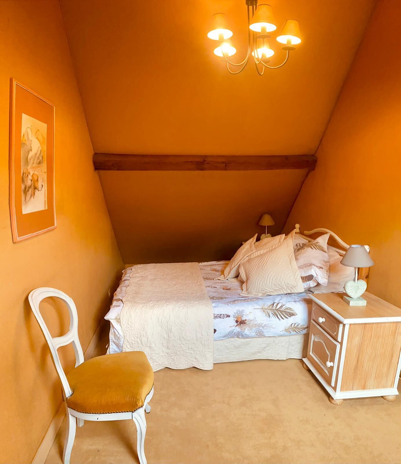 Une chambre mansardée chaleureuse, avec un plafond en pente, un lit recouvert d’une literie à motifs floraux, une lampe de chevet et une petite table de nuit.