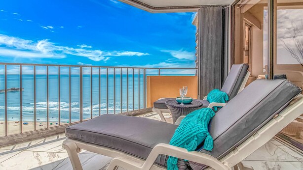 Een balkon met uitzicht op zee, een loungerstoel, een klein tafeltje met een glas en een decoratief kussen.