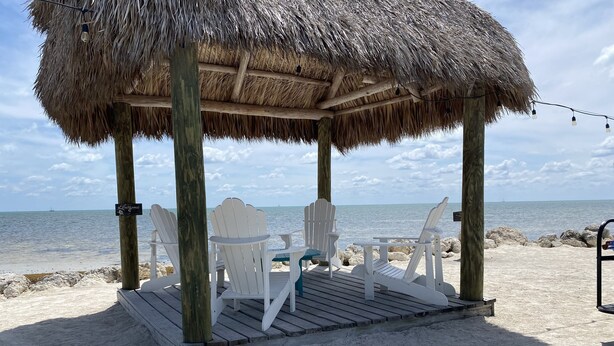 Un cabanon de plage avec des chaises Adirondack blanches et un toit de chaume, donnant sur l’océan.