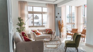 Een moderne woonkamer met een bank, fauteuil en salontafel, een groot raam met gordijnen en uitzicht op de stad.