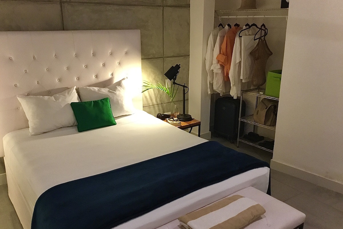 Ein ordentlich bezogenes Bett mit weißem Kopfteil, einem grünen Kissen und einer blauen Bettdecke. Ein Nachttisch mit Lampe und kleiner Pflanze steht daneben. Im Hintergrund ist ein Kleiderschrank mit Kleidung und einem Koffer zu sehen.