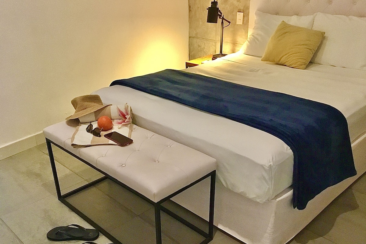 Ein ordentlich bezogenes Bett mit blauer Decke, einer Nachttischlampe und einem kleinen Tisch mit Hut und Sonnenbrille.