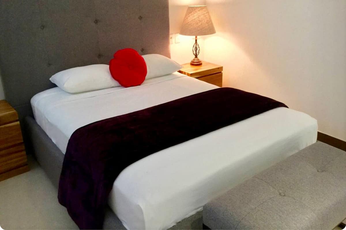Ein ordentlich bezogenes Bett mit grauem Kopfteil, weißer Bettwäsche, einem roten herzförmigen Kissen und einer dunkelroten Decke. Auf dem Nachttisch steht eine Lampe.