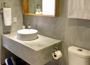 A bathroom with a round sink, a mirror, a toilet, and a roll of toilet paper on the floor. - Pilitas Vallarta Apartments (Puerto Vallarta)