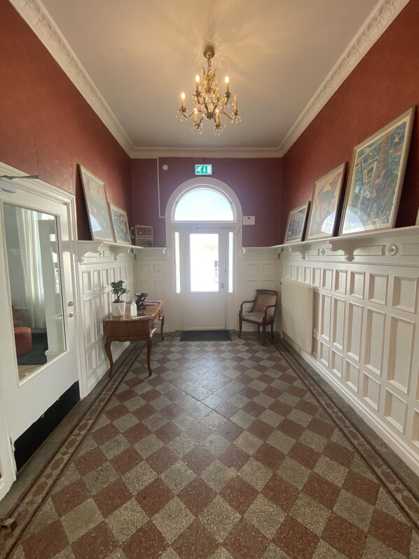 A hallway with checkered flooring, white paneling, a chandelier, framed artwork, and a door with a mirror. - Frövihotell (Frovi)