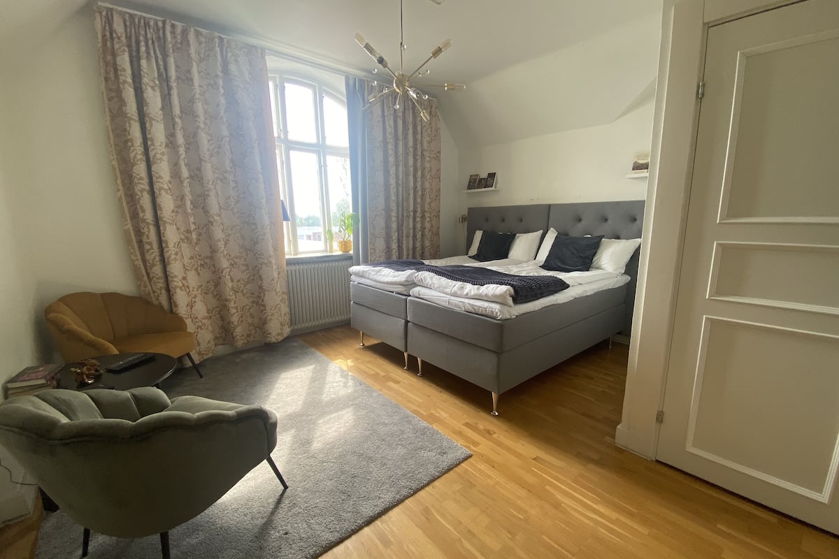 A bedroom with a bed, a chair, a radiator, a window with curtains, and a door.