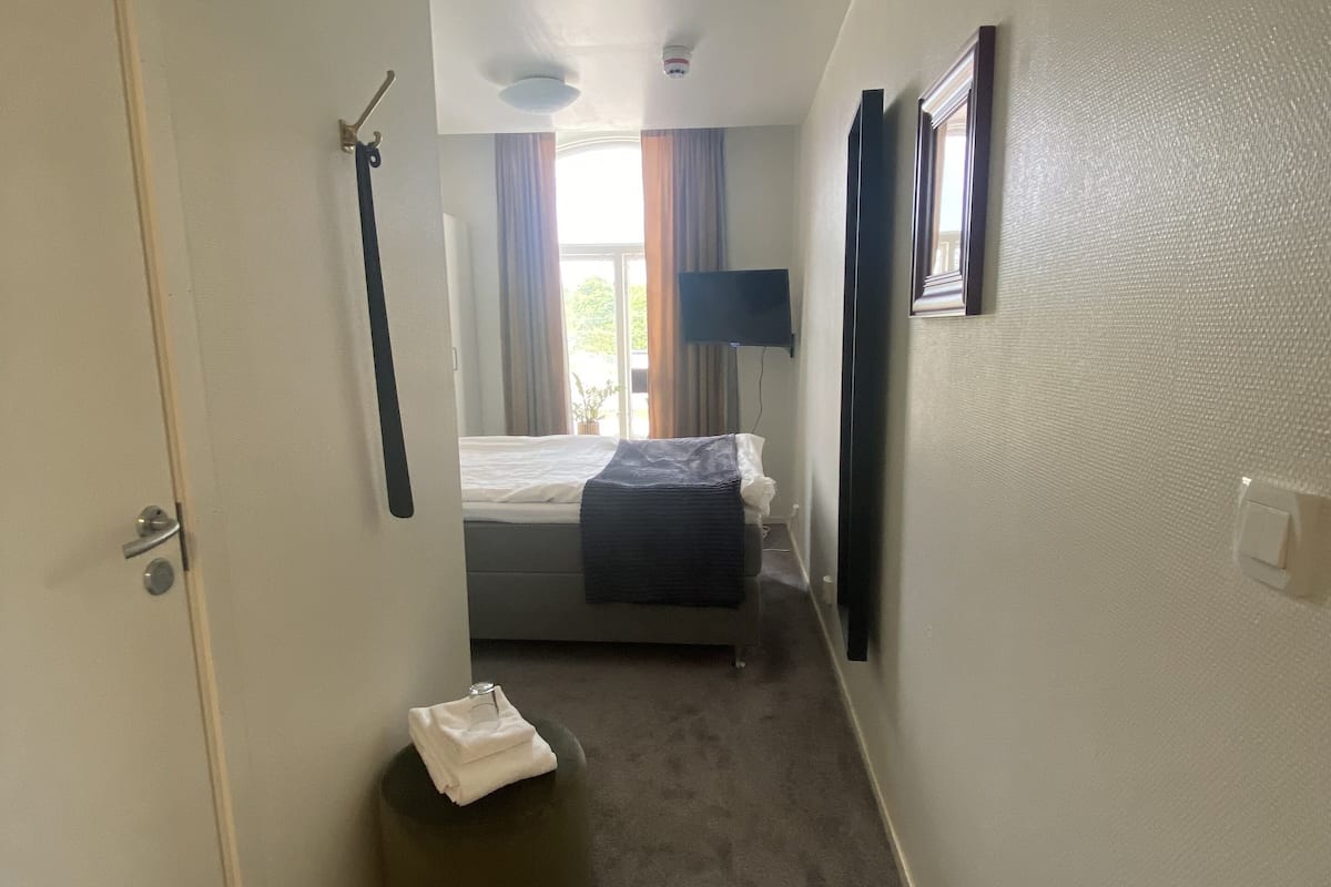 A narrow hallway leading to a bedroom with a bed, a small round ottoman, a flat-screen TV, and a ceiling light.
