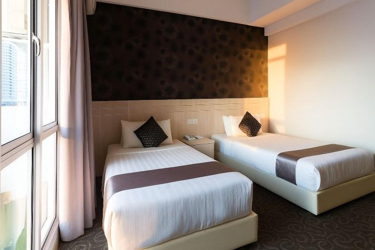 Quarto de hotel com duas camas, cabeceira escura e janela com cortinas.