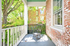 A porch with a rocking chair, a white railing, and a brick building in the background. - Hudson Mariner (Hudson)