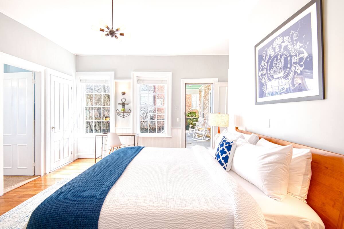A bedroom with a large bed, a chandelier, a framed picture, and a view through the window.
