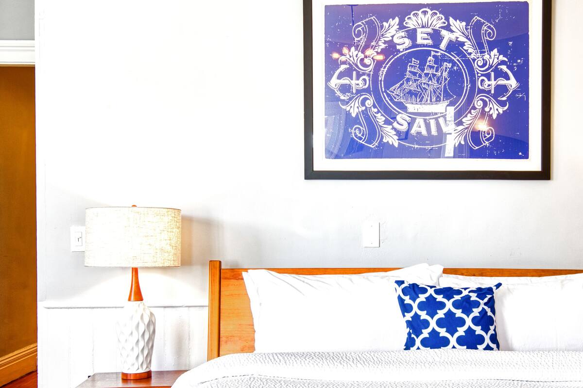 A neatly made bed with a blue and white bedspread, a framed picture on the wall, and a bedside table with a lamp and a decorative vase.