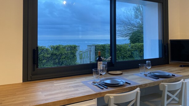 A dining area with a large window overlooking the sea, a wooden table set for two, and a modern kitchen visible to the right. - Maison Les Pieds Dans Leau Large vue sur mer 20 m de la Plage Wide Sea View (Loctudy)