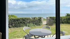 A view from inside a room to a patio with a round table and two chairs, overlooking a body of water and a grassy area with bushes. - Maison Les Pieds Dans Leau Large vue sur mer 20 m de la Plage Wide Sea View (Loctudy)