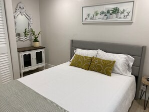 Een netjes ingerichte slaapkamer met een bed, een nachtkastje met een plant, een spiegel en een lijst met kunstwerk aan de muur.