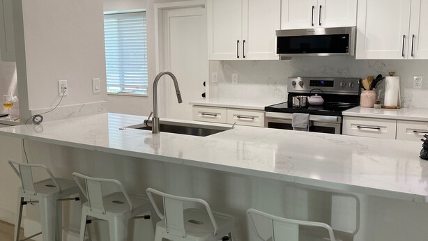 Een moderne keuken met witte kasten, een groot eiland met barstoelen, roestvrijstalen apparatuur en inbouwspots.