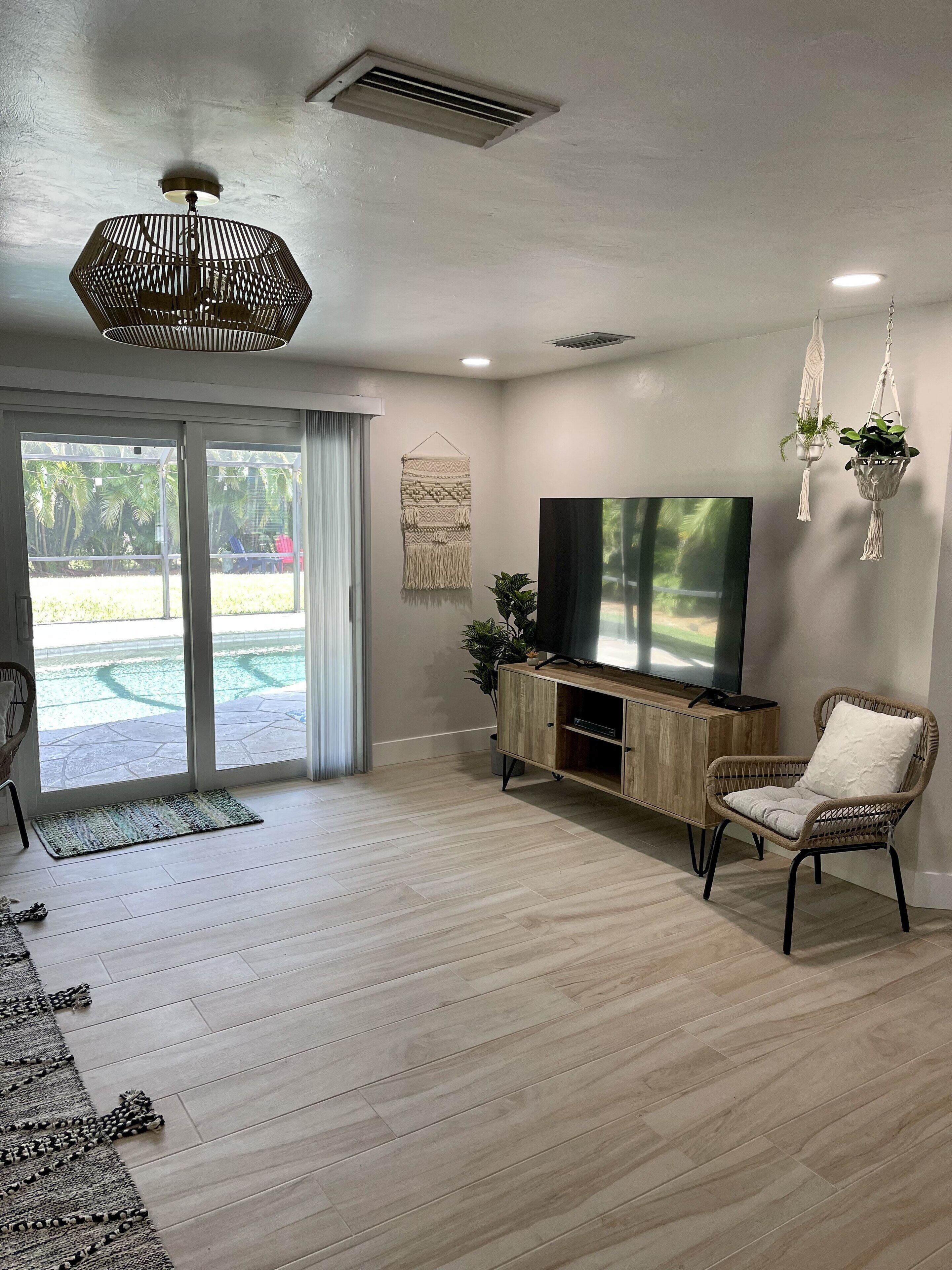 A modern living room with a flat-screen TV, wicker furniture, and a view of a pool through large sliding doors.