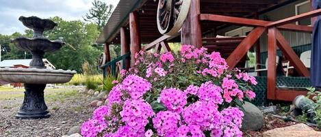 Un bâtiment rustique à la structure en bois, doté d’une grande roue et bordé d’un jardin où poussent des fleurs roses.