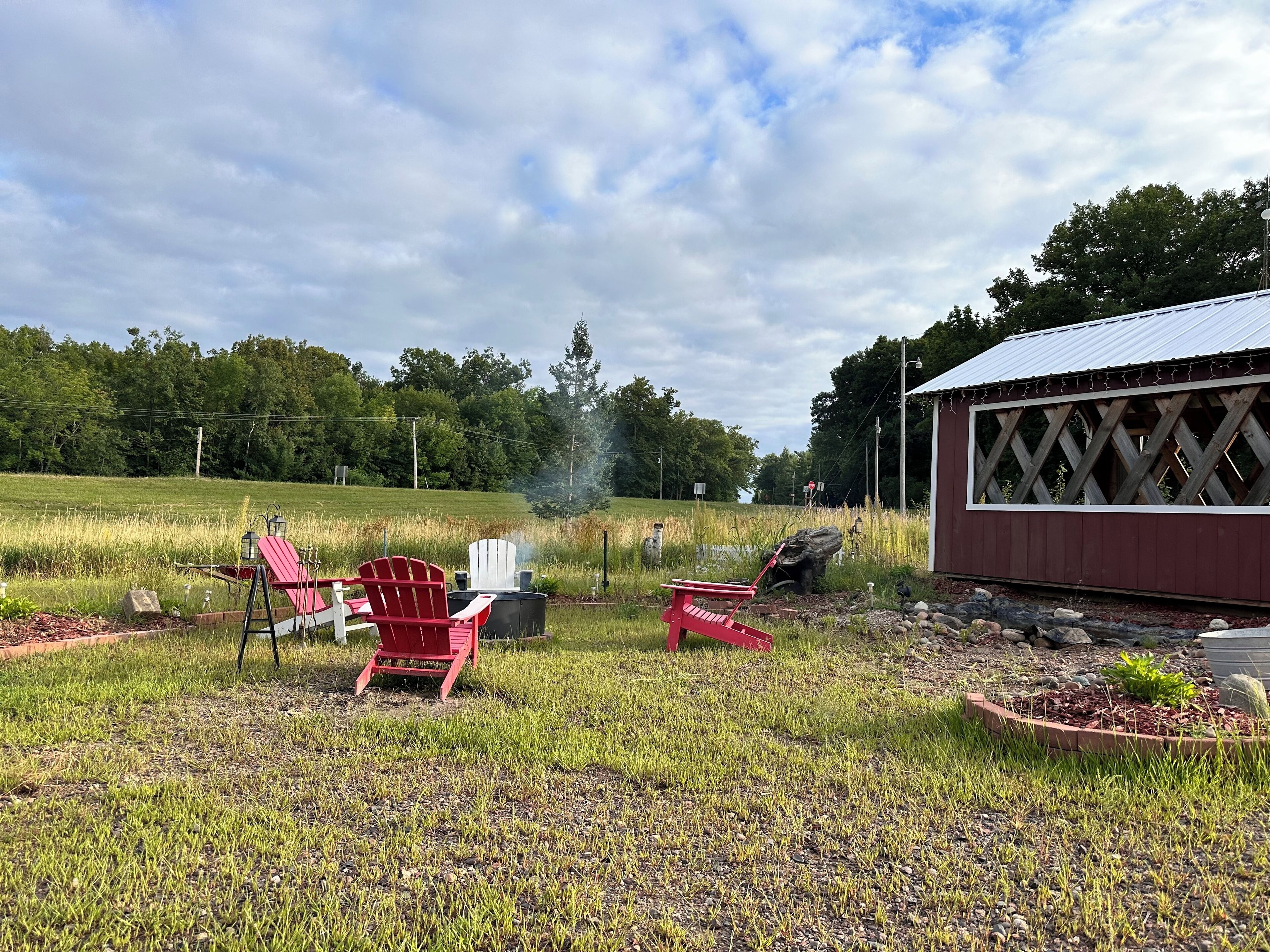 A rustic outdoor seating area with Adirondack chairs and a fire pit, surrounded by a grassy field and a wooden structure.
