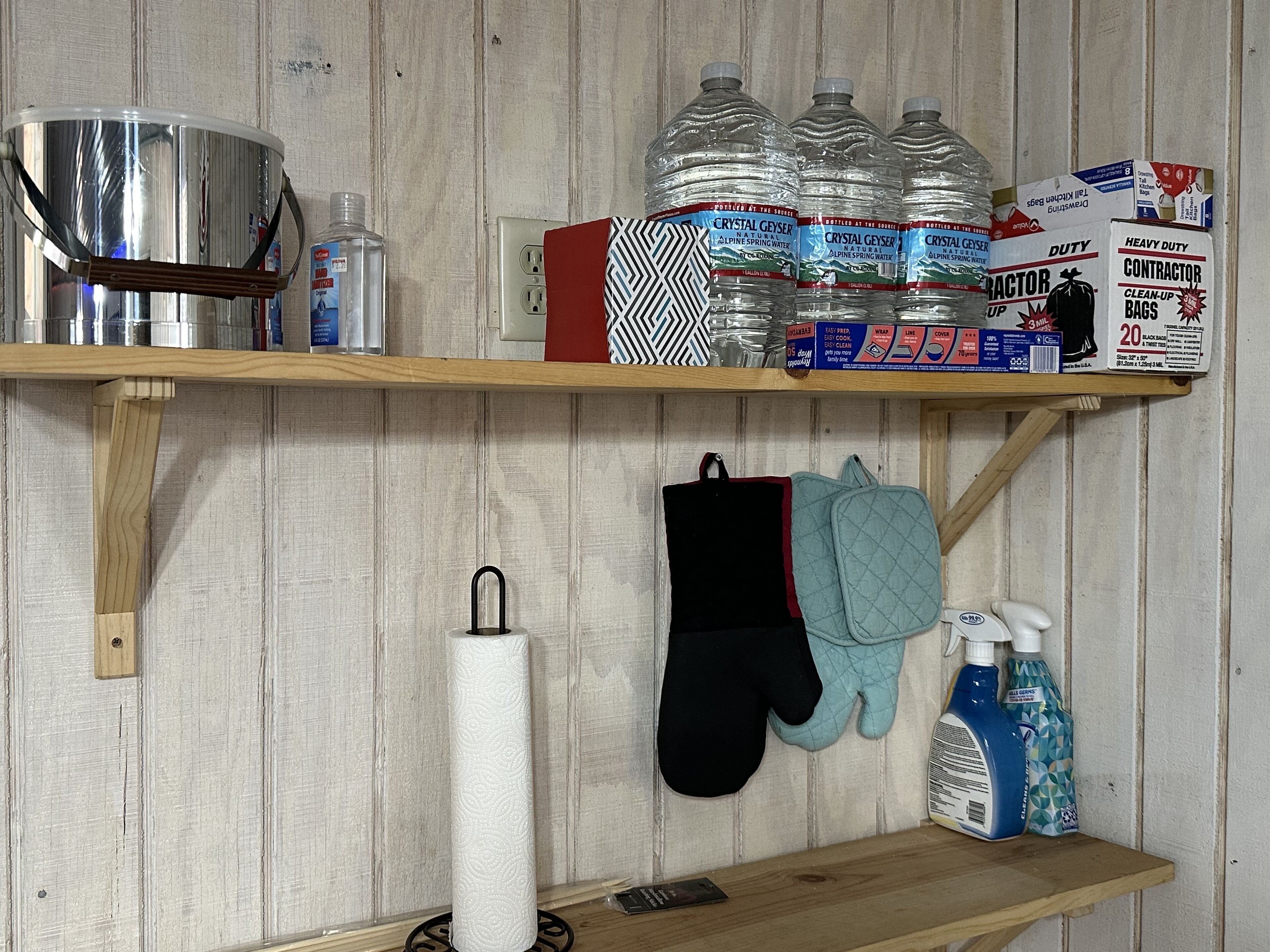 A kitchen shelf with a kettle, water bottles, cleaning supplies, and oven mitts.