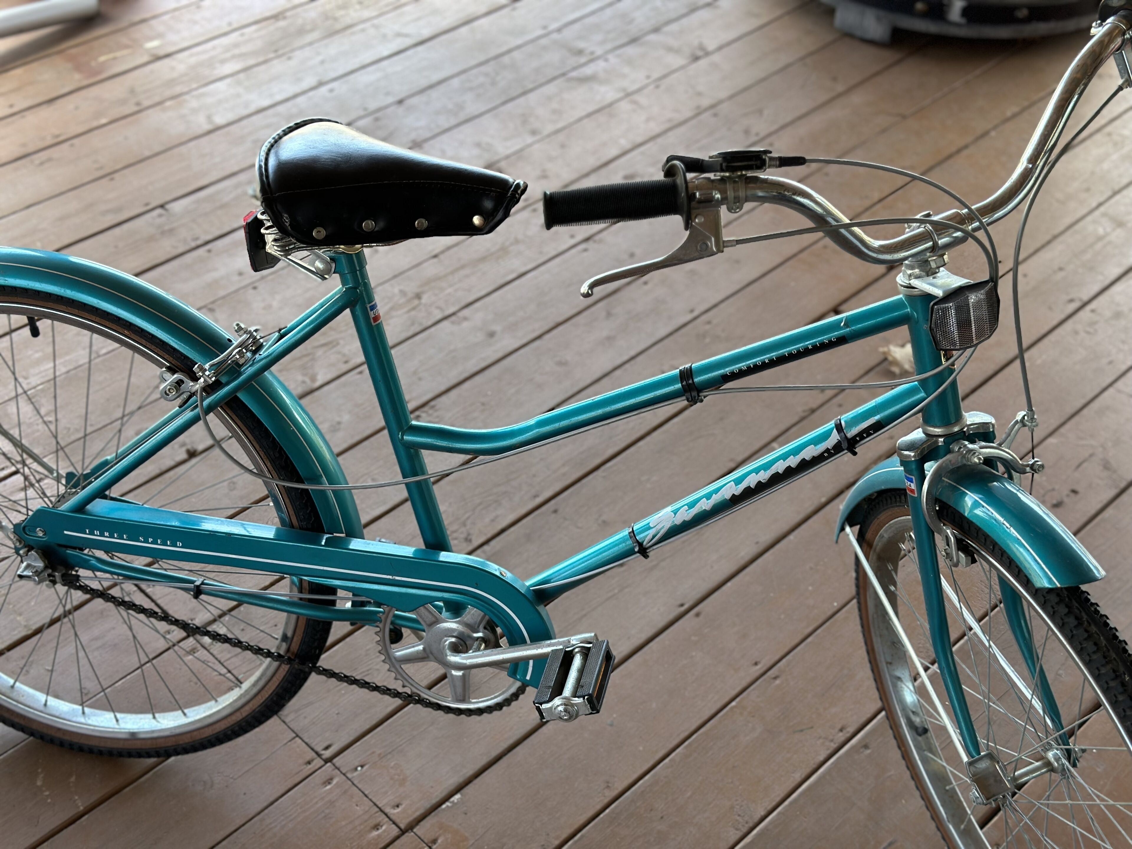 A teal bicycle with a black seat and a three-speed gear system, parked on a wooden deck.