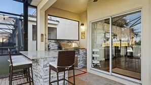 Un área de cocina al aire libre con una parrilla, taburetes de bar y una puerta corrediza de vidrio que da a un patio.