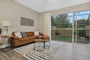 Una sala de estar moderna con un sofá de cuero marrón, dos mesitas auxiliares, un tapete blanco y negro y un gran ventanal corredero que da a un área exterior con piscina.