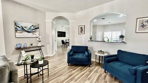 A spacious living room with a wooden floor, a blue armchair, a sofa, and a dining area in the background. - Large 6BR w/ FREE water park and private pool (Kissimmee)