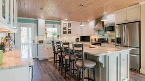 A modern kitchen with a central island, bar stools, stainless steel appliances, and a wooden ceiling.