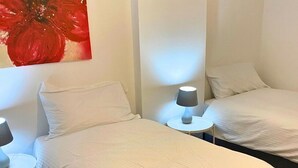 Ein Hotelzimmer mit zwei Einzelbetten, weißer Bettwäsche, einem runden Beistelltisch mit Lampe und einem roten abstrakten Gemälde an der Wand.