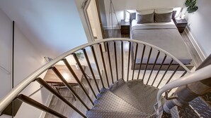 Eine Wendeltreppe mit Metallgeländern führt zu einem Schlafzimmer, das ein Bett, ein Nachttisch, eine Lampe und eine Pflanze enthält.