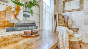 Une table de salle à manger en bois, ornée d’un chemin de table en dentelle, un vase avec des fleurs et une assiette de nourriture. Des livres sont empilés sur la table, et une chaise est recouverte d’un plaid tricoté.