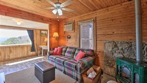 A cozy cabin interior with a plaid sofa, a fireplace, a ceiling fan, and a large window offering a view. - Secluded Ridgetop Hideaway w/ Valley Views! (Menlo)