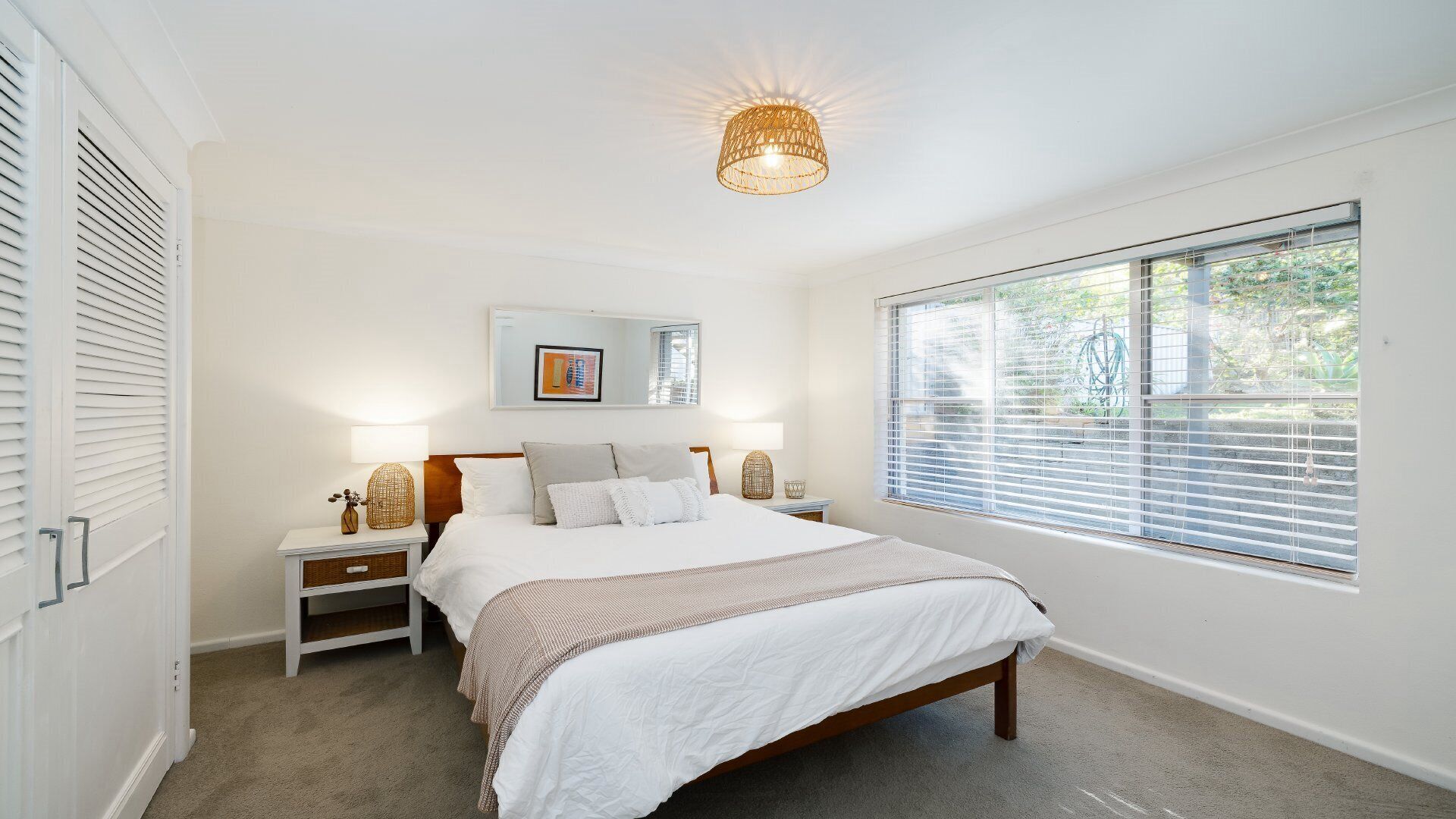 A bedroom with a large bed, two bedside tables, a wall-mounted artwork, a window with blinds, and a ceiling light fixture.