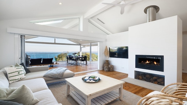 A modern living room with a fireplace, a sofa, a dining area, and a view of the sea.