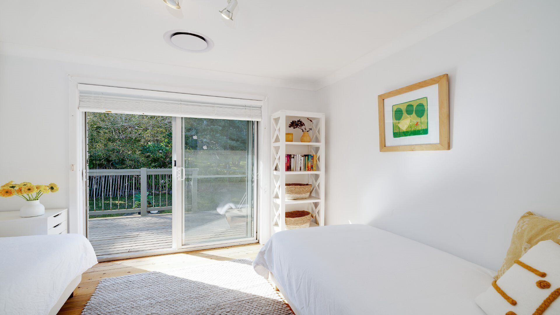A bedroom with a sliding glass door leading to a balcony, a white bed, a bedside table with a vase of flowers, a white bookshelf, and a framed picture on the wall.