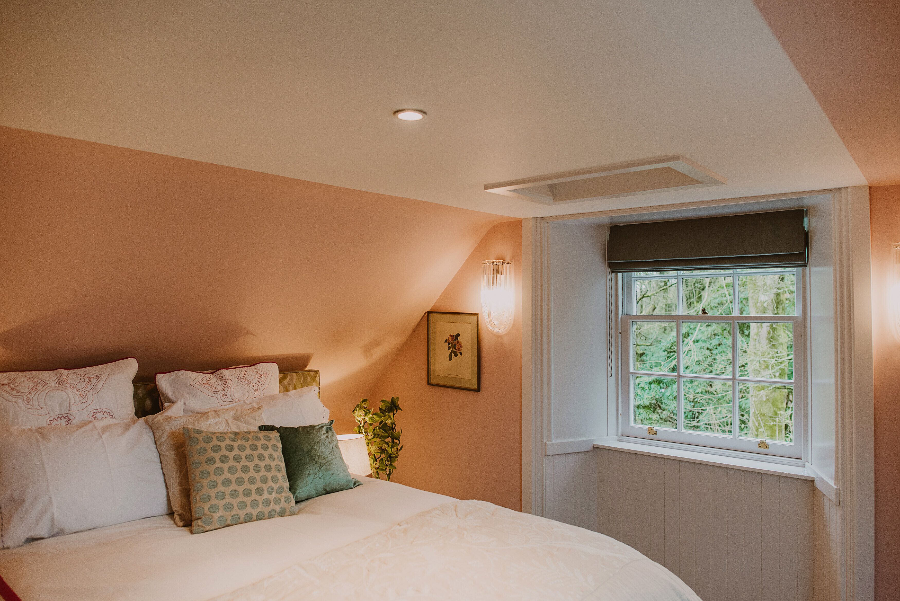 A bedroom with a bed, pillows, a wall-mounted light, a window with a view of greenery, and a ceiling vent.