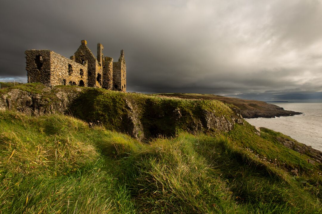 Un château de pierre historique, perché sur une falaise verdoyante qui surplombe la mer, sous un ciel nuageux.
