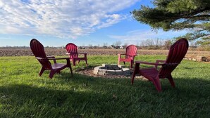 A group of Adirondack chairs arranged around a fire pit in a grassy field with a clear sky. - Soldiers Home (West Lafayette)