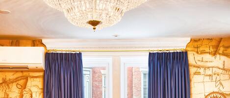 A room with a large window, two blue chairs, a round table, a chandelier, and a map wall.