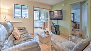 Un salon comprenant un canapé, un fauteuil et un téléviseur fixé au mur. On y trouve également une cheminée et une porte qui donne sur une autre pièce.