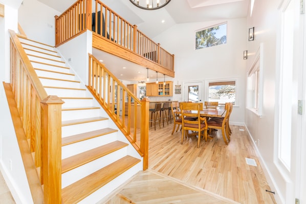 A spacious interior with a wooden staircase leading to a mezzanine, a dining area with wooden furniture, and a view through a window.