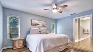 Ein Schlafzimmer mit Bett, Deckenventilator, Fenster mit Jalousien, Nachttisch und Spiegel.