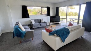 A modern living room with a grey sofa, a white sofa, and a grey armchair. There is a flat-screen TV on a wooden stand, a small wooden table, and a large window with black curtains. - Coopers Haven Beachfront House (Coopers Beach)