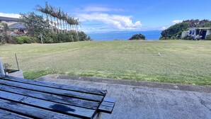 A patio with a wooden table and chairs, overlooking a grassy area with a view of the ocean and distant hills. - Coopers Haven Beachfront House (Coopers Beach)