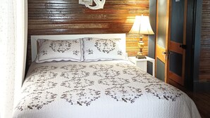 Een slaapkamer met een houten plafond, een bed met een bloemenbedekking, een decoratieve ankerhanger aan de muur, en een nachtkastje met een lamp.