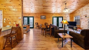 Een houten chalet van binnen met een eethoek, een bank, een televisie en een plafondventilator.
