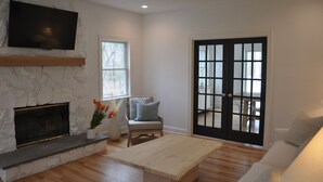 Un salón con chimenea de piedra, un televisor de pantalla plana montado encima, un sillón cómodo, un sofá y un amplio ventanal corredero.