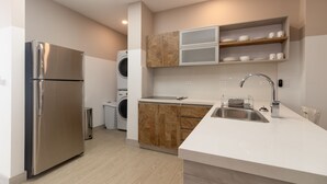 A modern kitchen with a stainless steel refrigerator, built-in washing machine, wooden cabinets, and a white countertop.